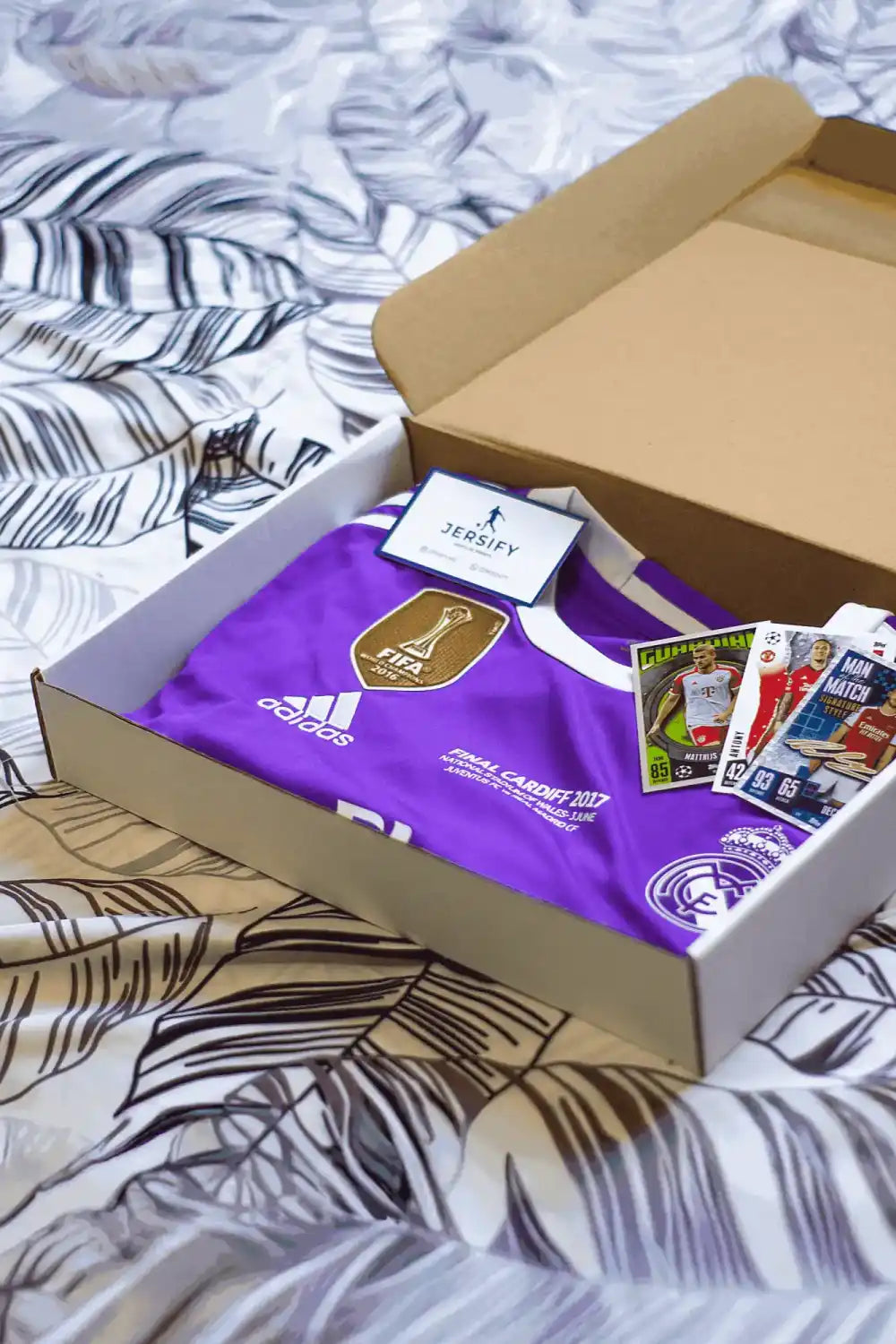 Camiseta de fútbol/soccer morada en una caja de cartón abierta con algunas tarjetas de intercambio visibles.